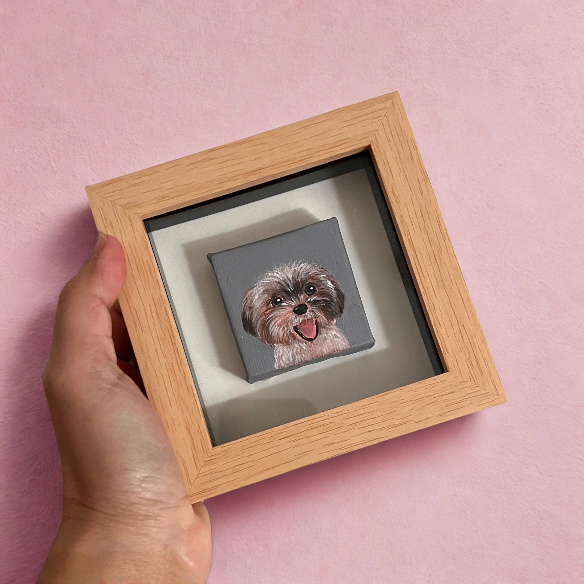 Hand holding a small framed artwork of a dog on a pink background