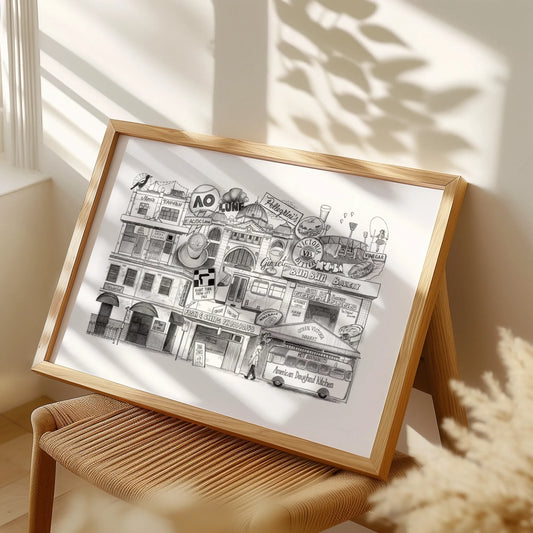 Framed black and white illustration of a city street scene on a white background, placed on a wooden stool.